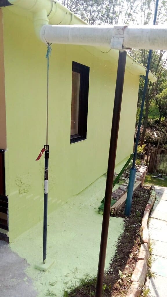 Hliníková tyč s hákem u domu, zelené zdivo, okno.