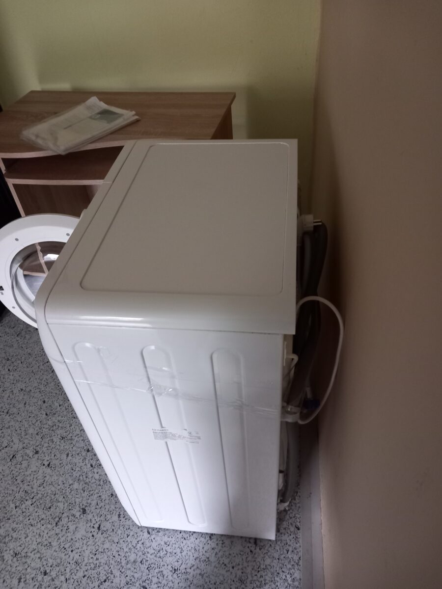 Pračka stojí u zdi v domácím prostředí, vedle ní je malý nábytek a na něm jsou noviny nebo časopisy.